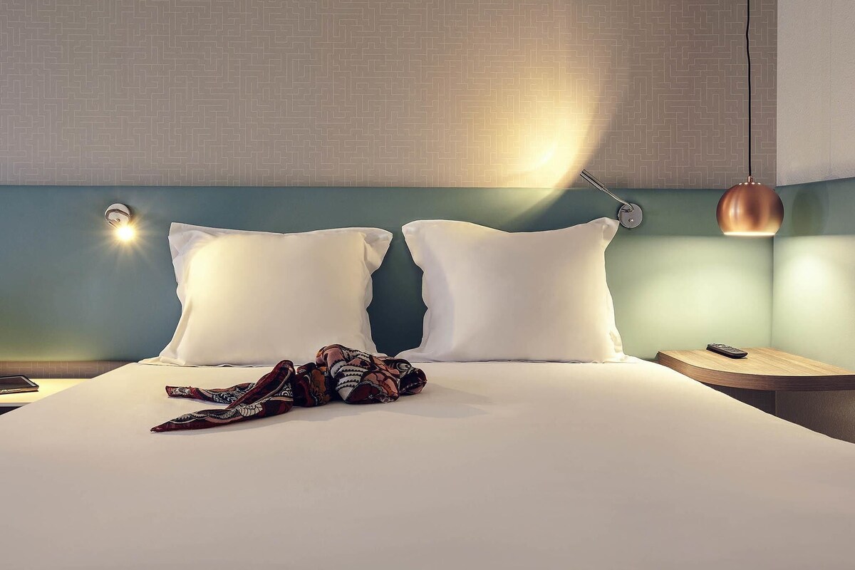 A neatly made bed with pillows, a patterned blanket, and a remote control on a wooden nightstand.