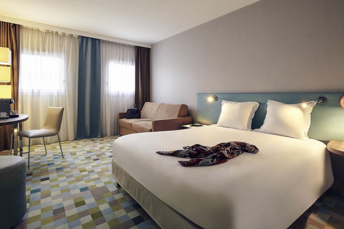 A hotel room with a large bed, a desk with a chair, a sofa, and a patterned carpet.