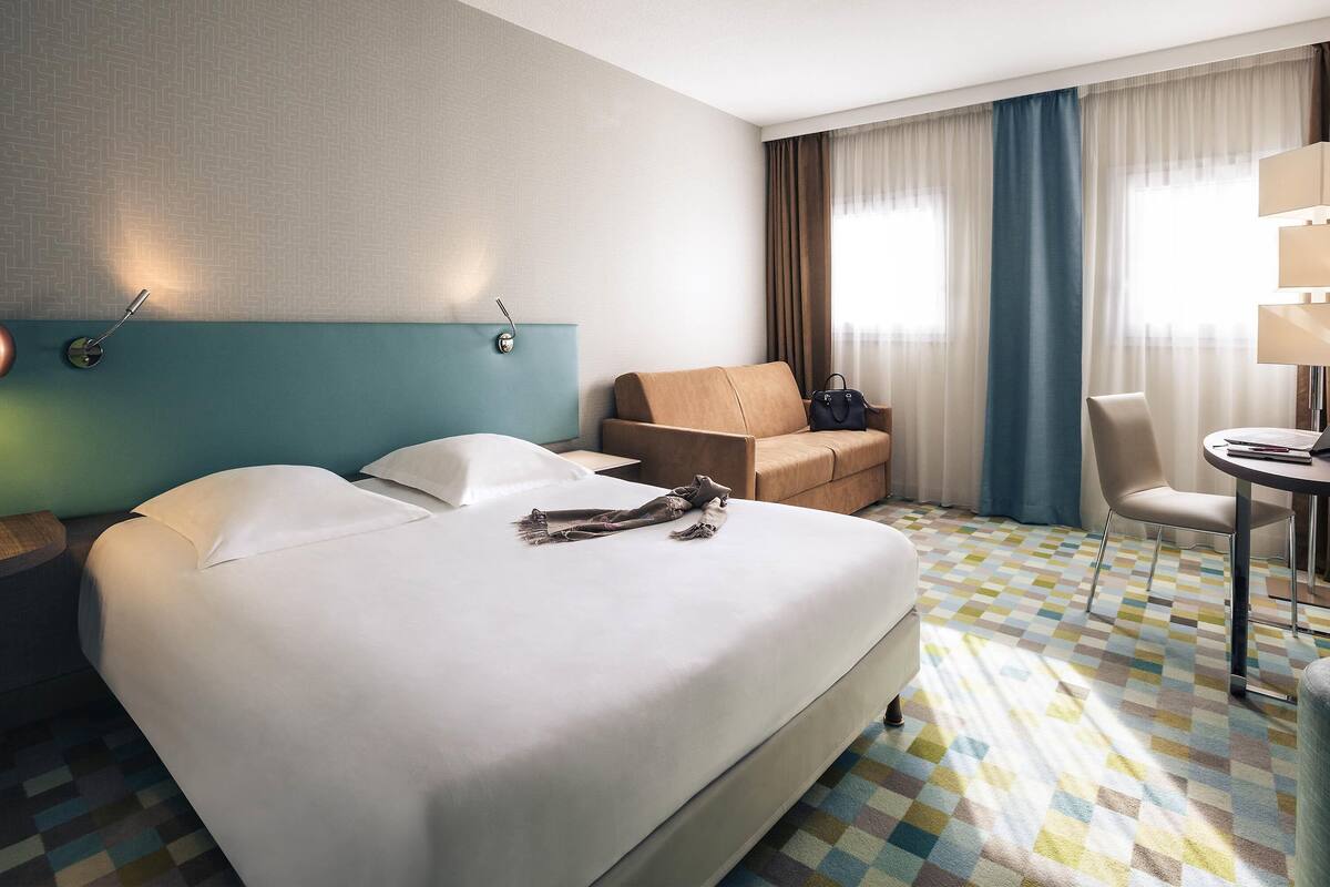A hotel room with a large bed, a sofa, a desk with a chair, and a lamp.