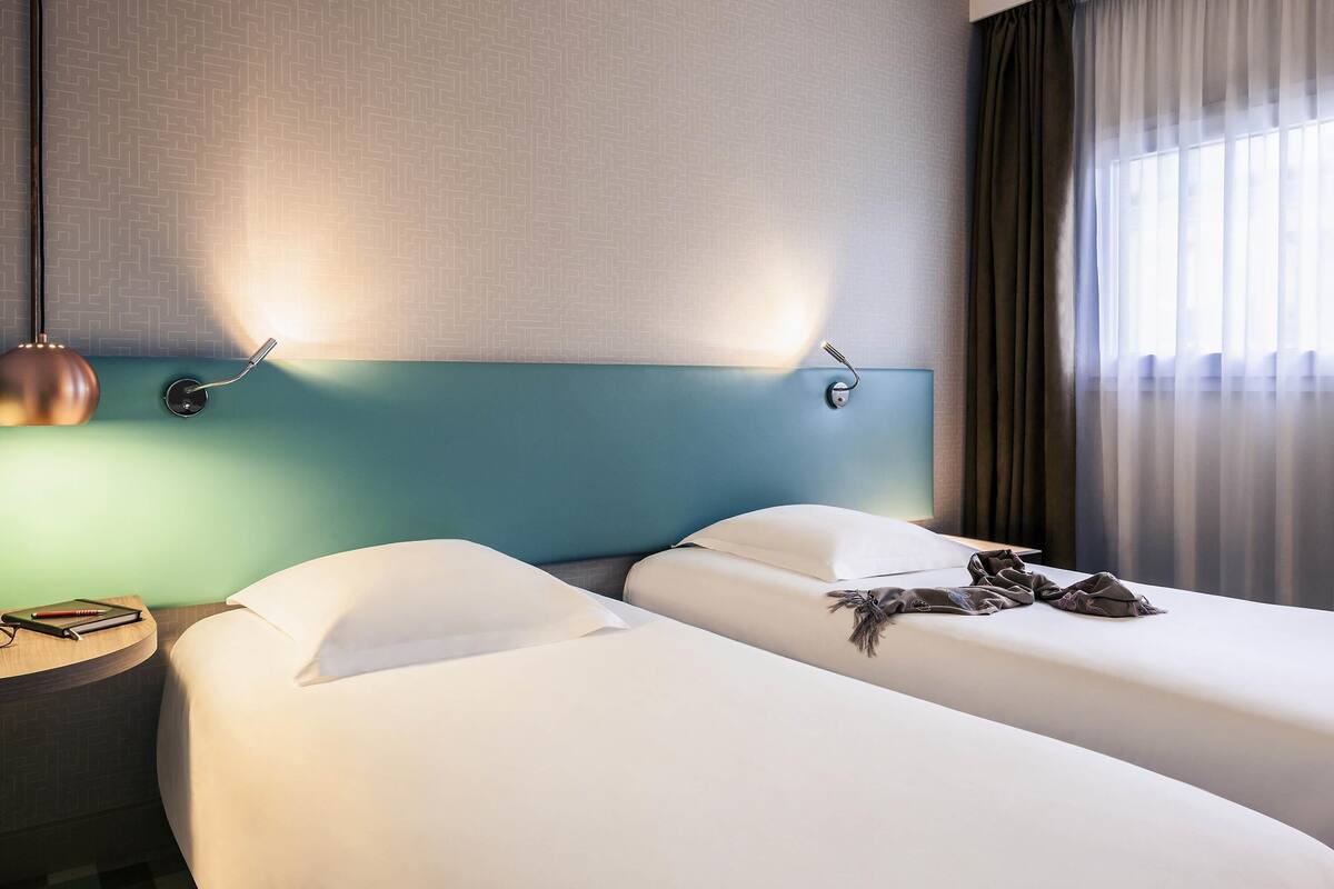 A hotel room with two beds, a nightstand, a lamp, and a window with curtains.