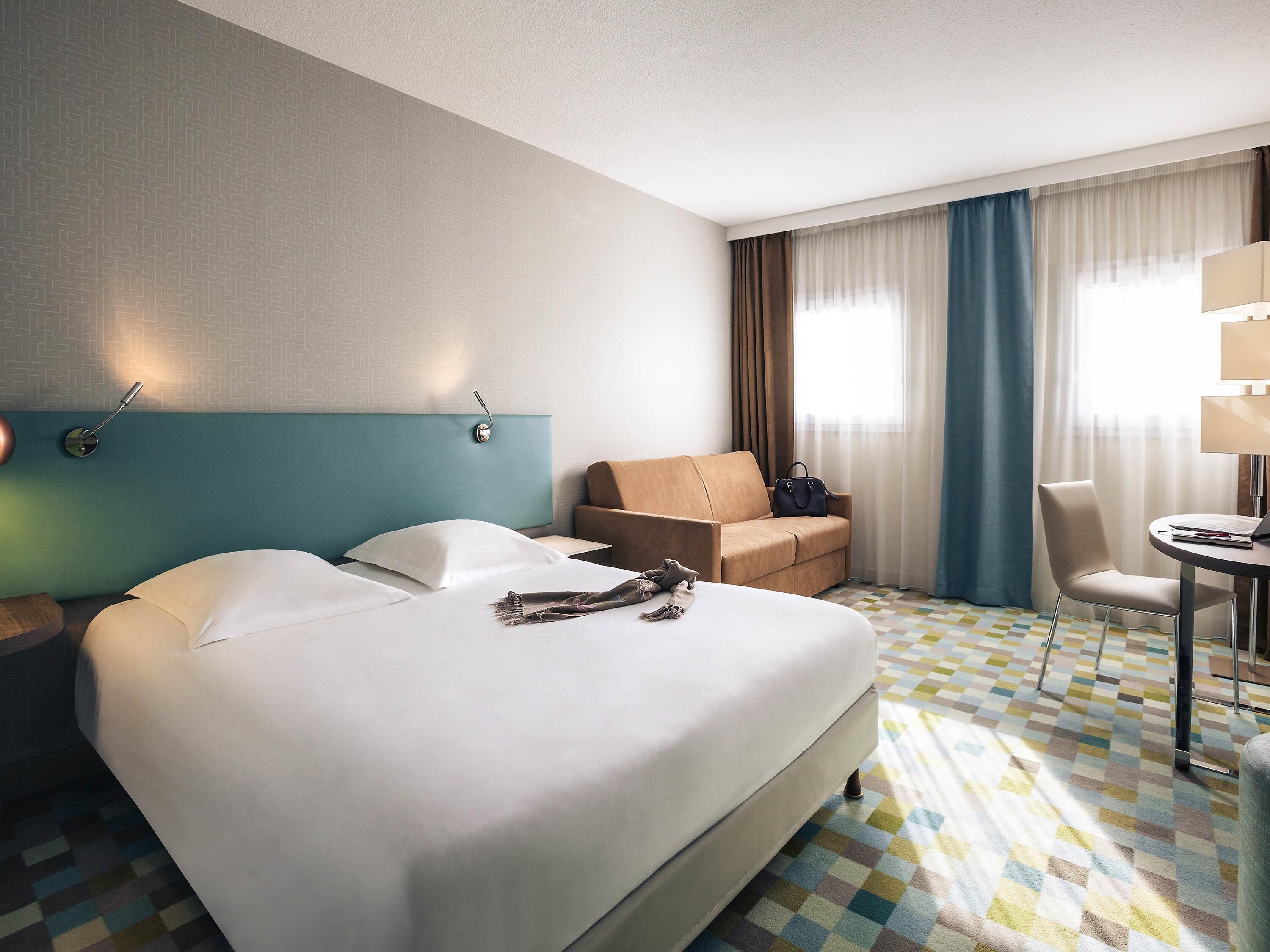 A hotel room with a large bed, a sofa, a desk with a chair, a lamp, and a window with curtains.