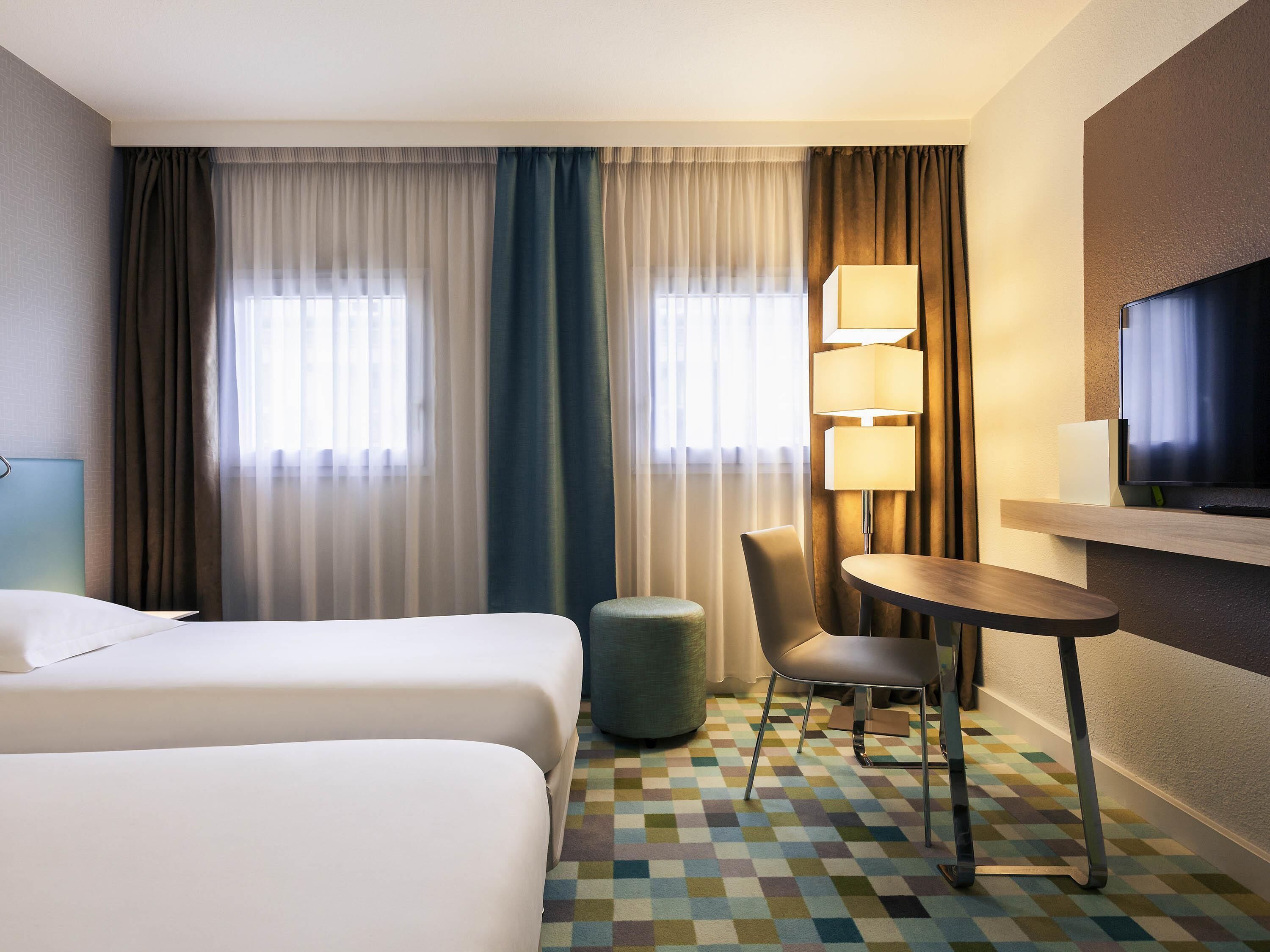 A hotel room with two beds, a desk, a chair, a TV, and a lamp.
