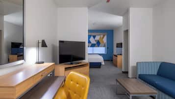 A modern hotel room with a wooden desk, a yellow chair, a blue sofa, a flat-screen TV, a bed with a geometric-patterned headboard, and a large mirror.