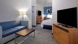 A hotel room with a blue sofa, a wooden desk, a TV, a bed, and a desk lamp. - Holiday Inn Express Hotel & Suites Henderson by IHG (Henderson)