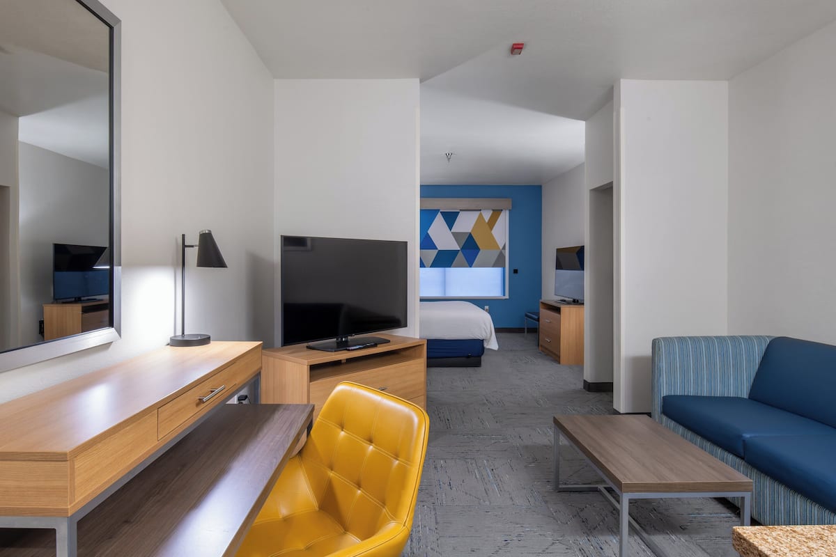 A modern hotel room with a wooden desk, a yellow chair, a blue sofa, a flat-screen TV, a bed with a geometric-patterned headboard, and a large mirror.