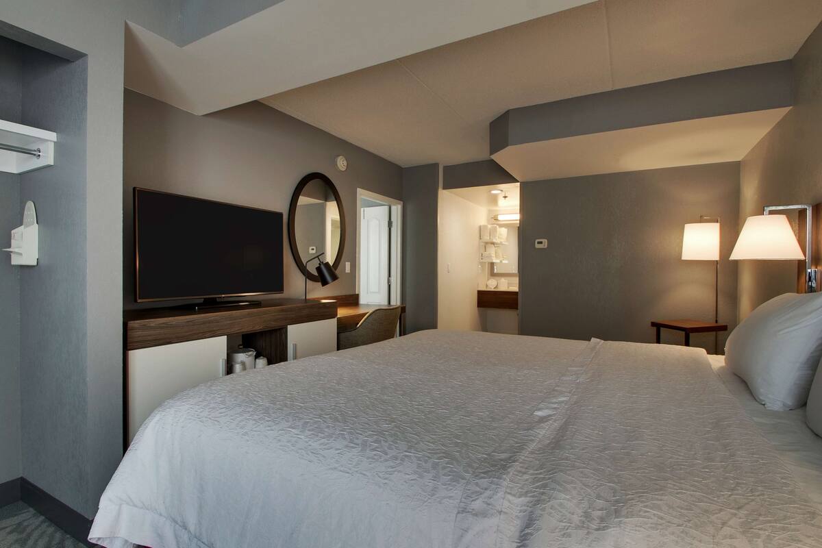 Habitación de hotel con una cama grande, televisión de pantalla plana, escritorio con silla, espejo y lámpara.