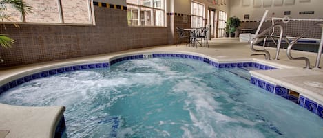 A hot tub with a blue and white tile border, indoor seating area with chairs, and a brick wall with windows.