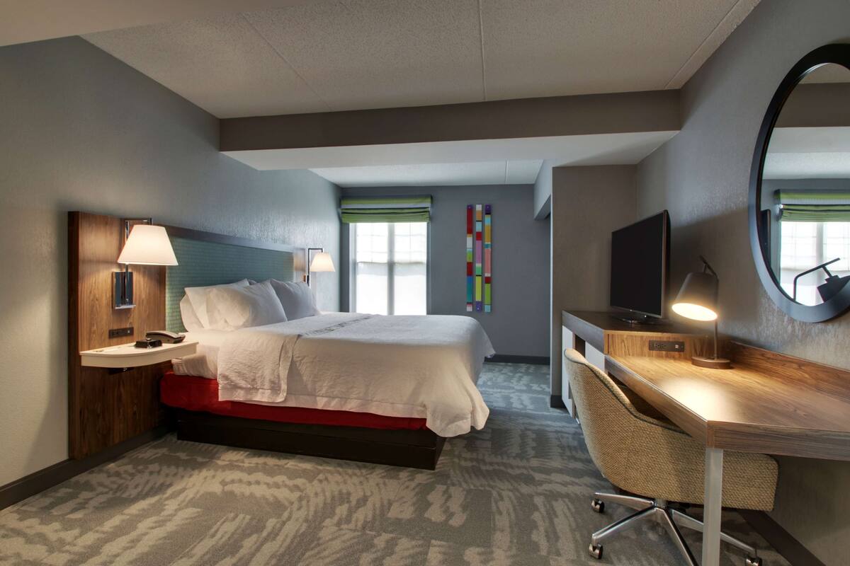 Habitación de hotel con cama, mesita de noche, lámpara, escritorio, silla, televisión y un espejo grande.