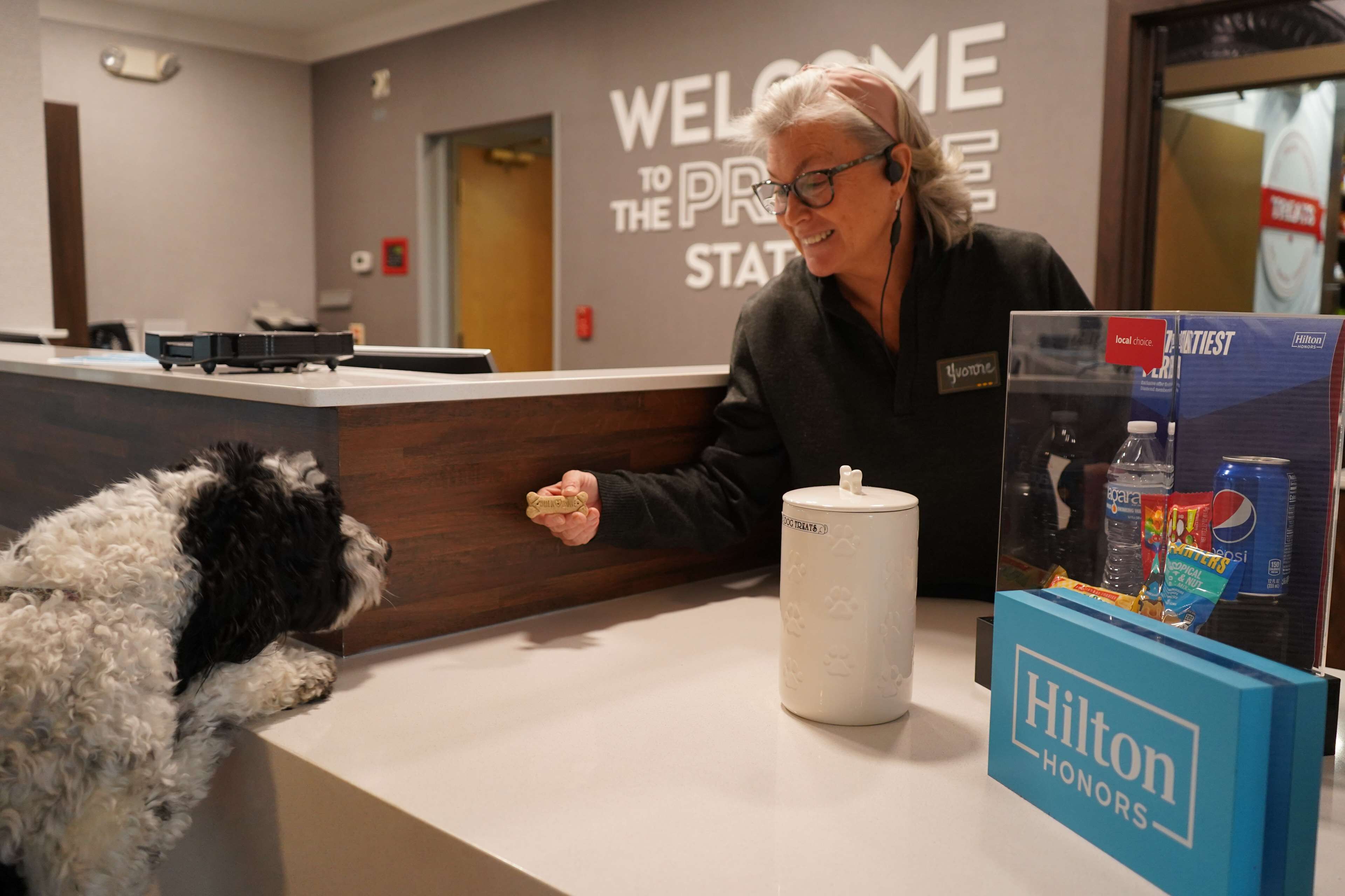 A hotel reception area with a 'Welcome to the Premier State' sign, a white pet-friendly reception desk, and a Hilton Honors sign.