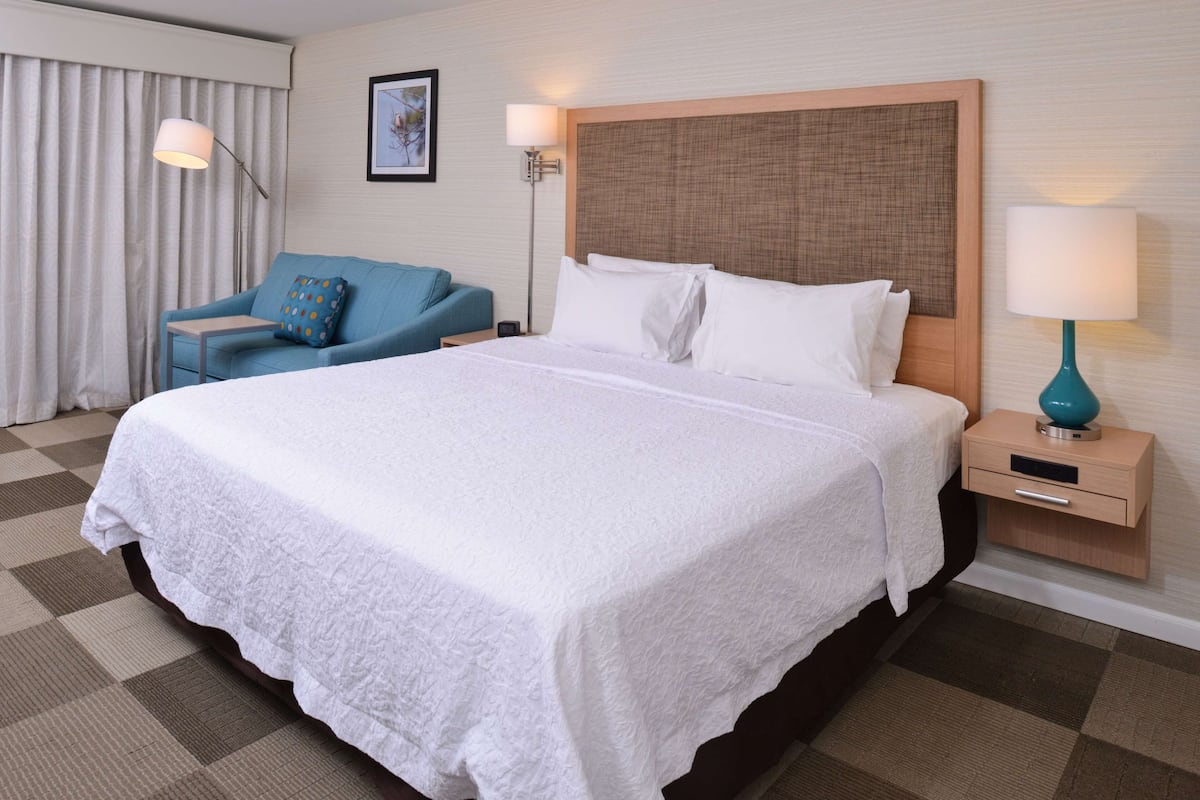 A hotel room with a large bed, a blue armchair, a nightstand with a lamp, and a wall-mounted lamp.