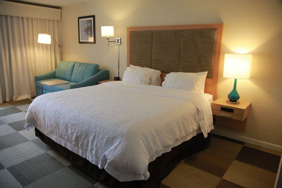 A hotel room with a large bed, a small sofa, a nightstand with a lamp, and a window with curtains.