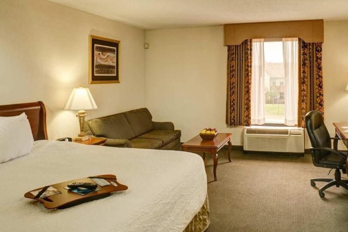 A hotel room with a bed, a sofa, a desk, a chair, a lamp, a window with curtains, and a framed picture on the wall.