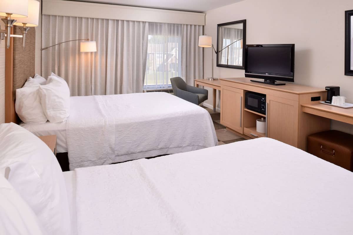 A hotel room with two beds, a desk with a television, a chair, and a minibar.