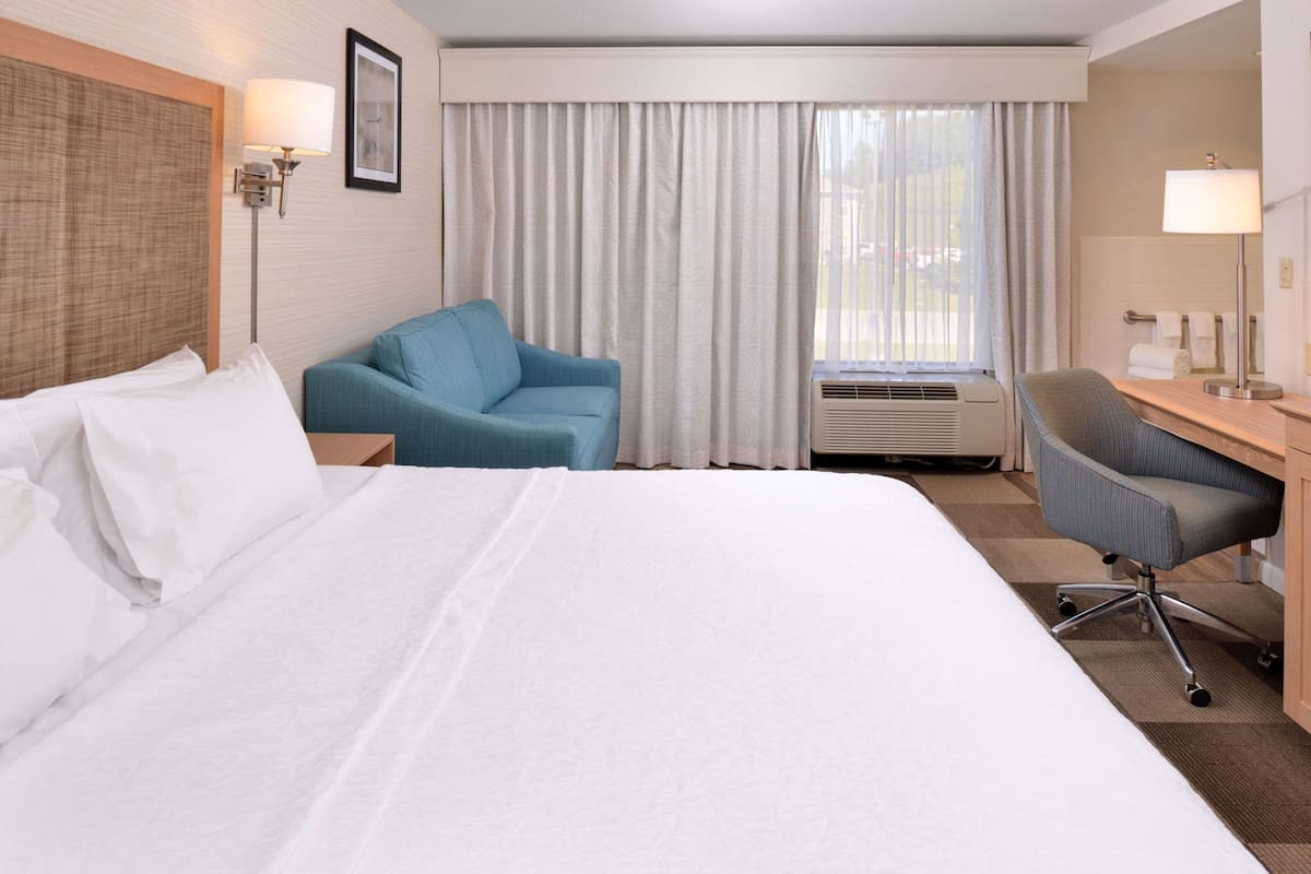 A hotel room with a large bed, a blue armchair, a desk with a chair, a window with curtains, and a wall-mounted lamp.