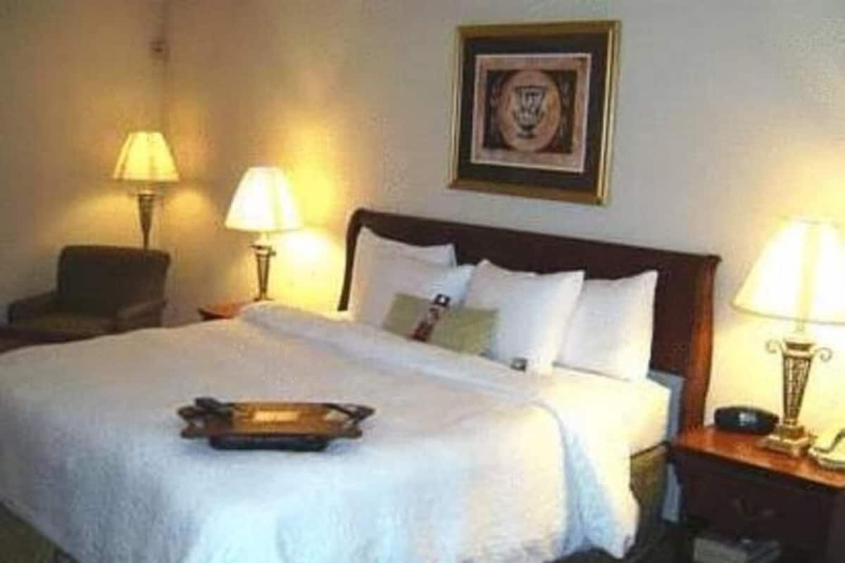 A hotel room with a bed, two bedside lamps, a chair, and a framed picture on the wall.