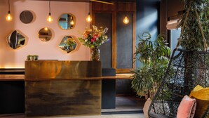 A reception area with a wooden reception desk, multiple mirrors, hanging light bulbs, a potted plant, and a wicker chair with cushions.