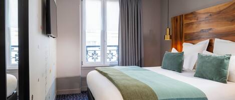 A hotel room with a bed, a window with curtains, a wooden headboard, and a wall-mounted TV.