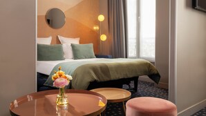 A hotel room with a bed, a round table with a vase of flowers, a small stool, and a large window with curtains.