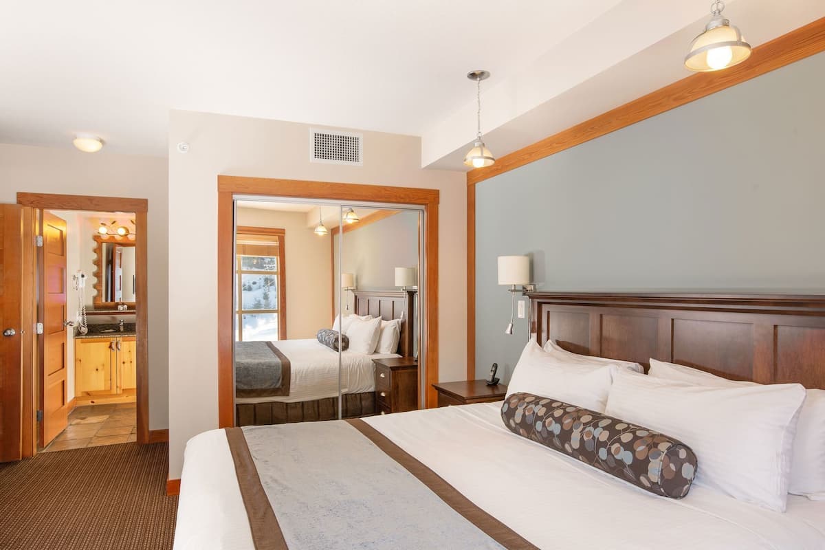 A hotel room with a large bed, a wooden headboard, a nightstand with a lamp, a mirror, and a bathroom visible through an open door.
