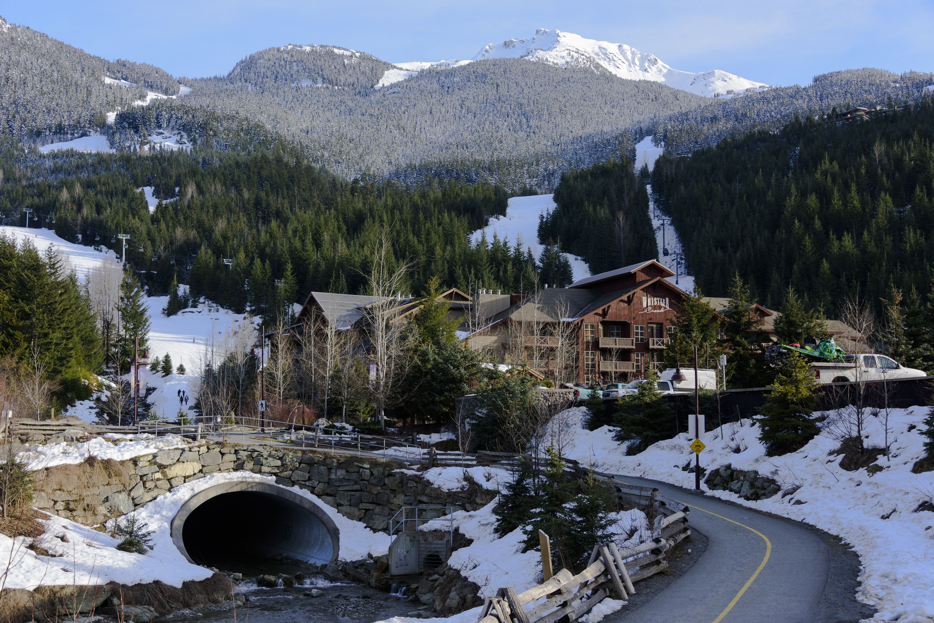 A mountain lodge with a stone bridge, a tunnel, and a ski slope in the background.