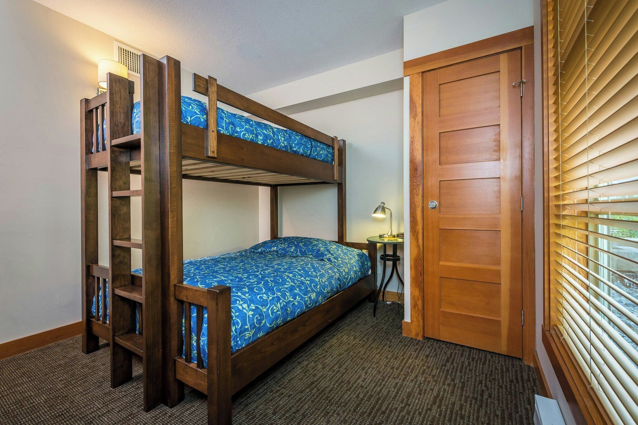 A bunk bed with a ladder, a small table with a lamp, and a wooden door.