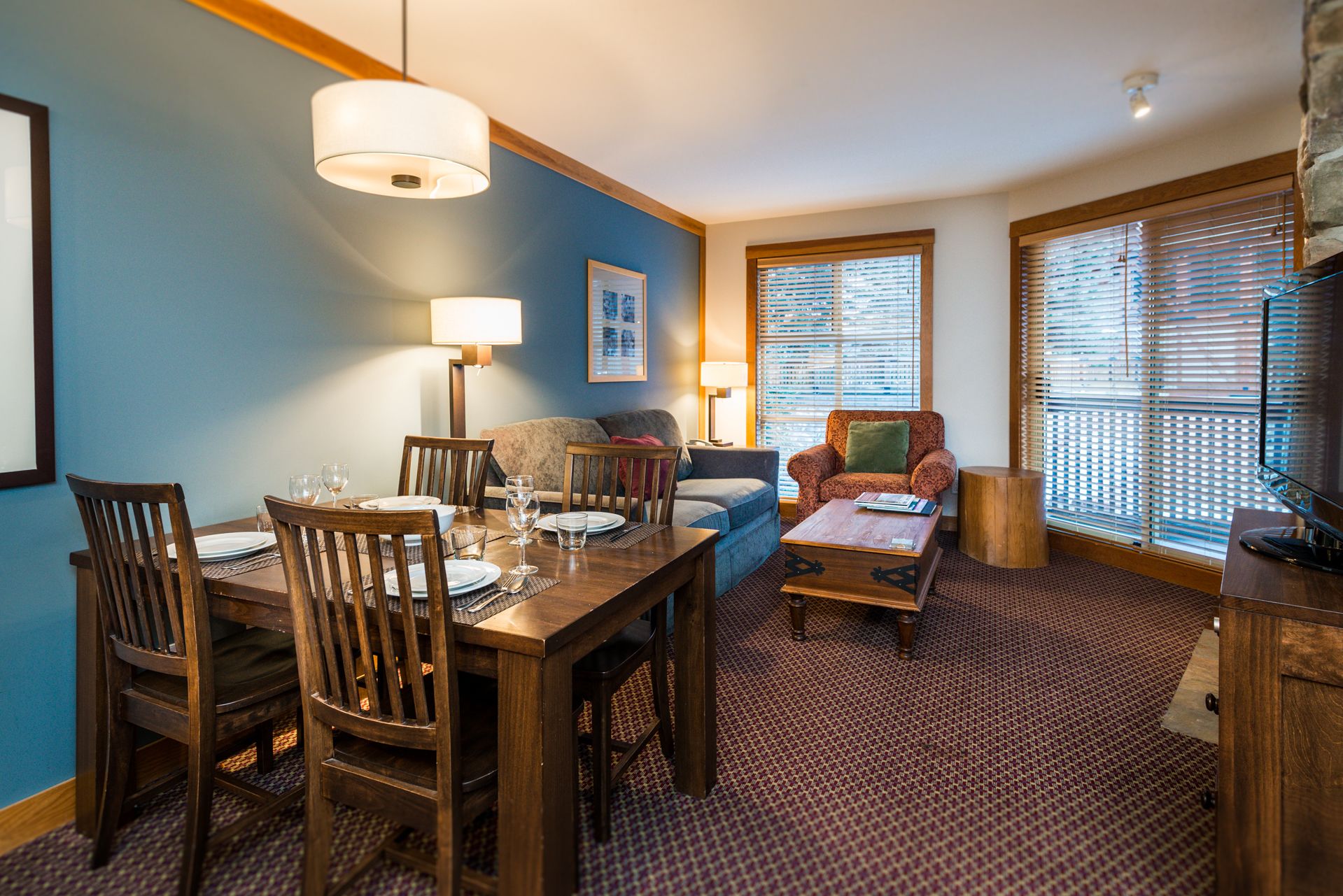 A dining area with a wooden table and chairs, a sofa, and a coffee table. There are two windows with blinds and a lamp on the wall.