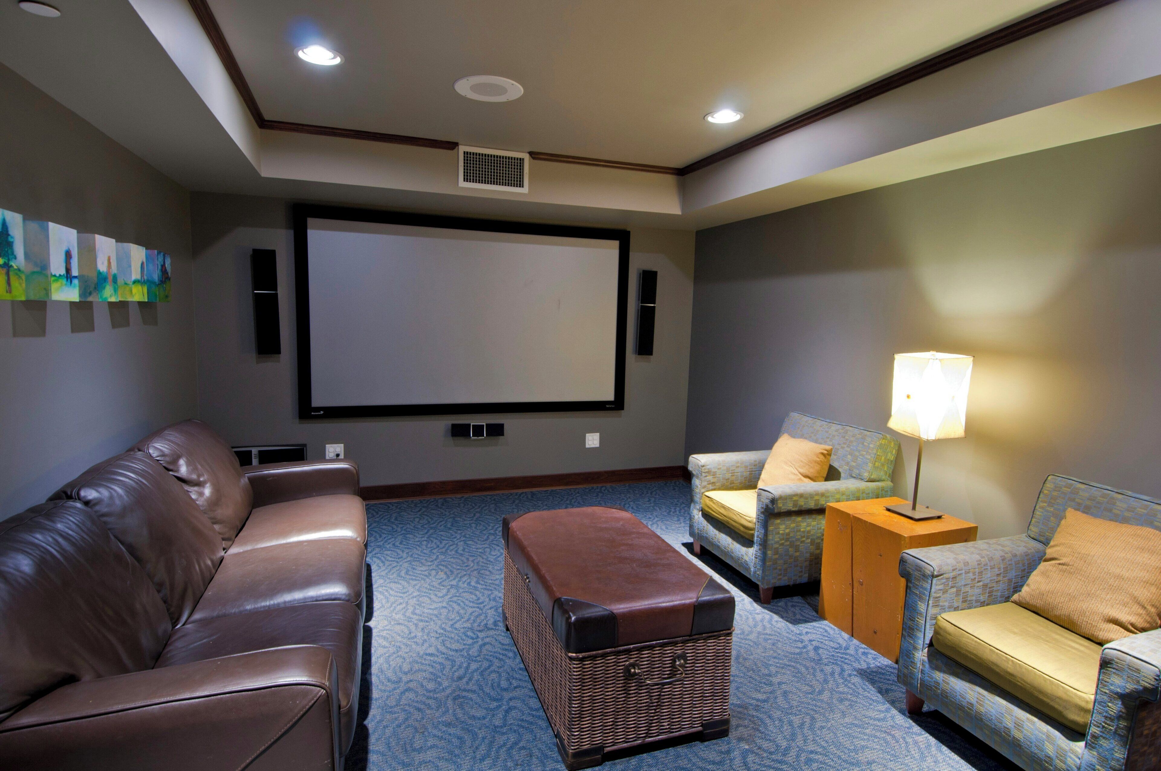 A home theater room with a brown leather couch, two patterned armchairs, a small wooden side table with a lamp, and a large screen.