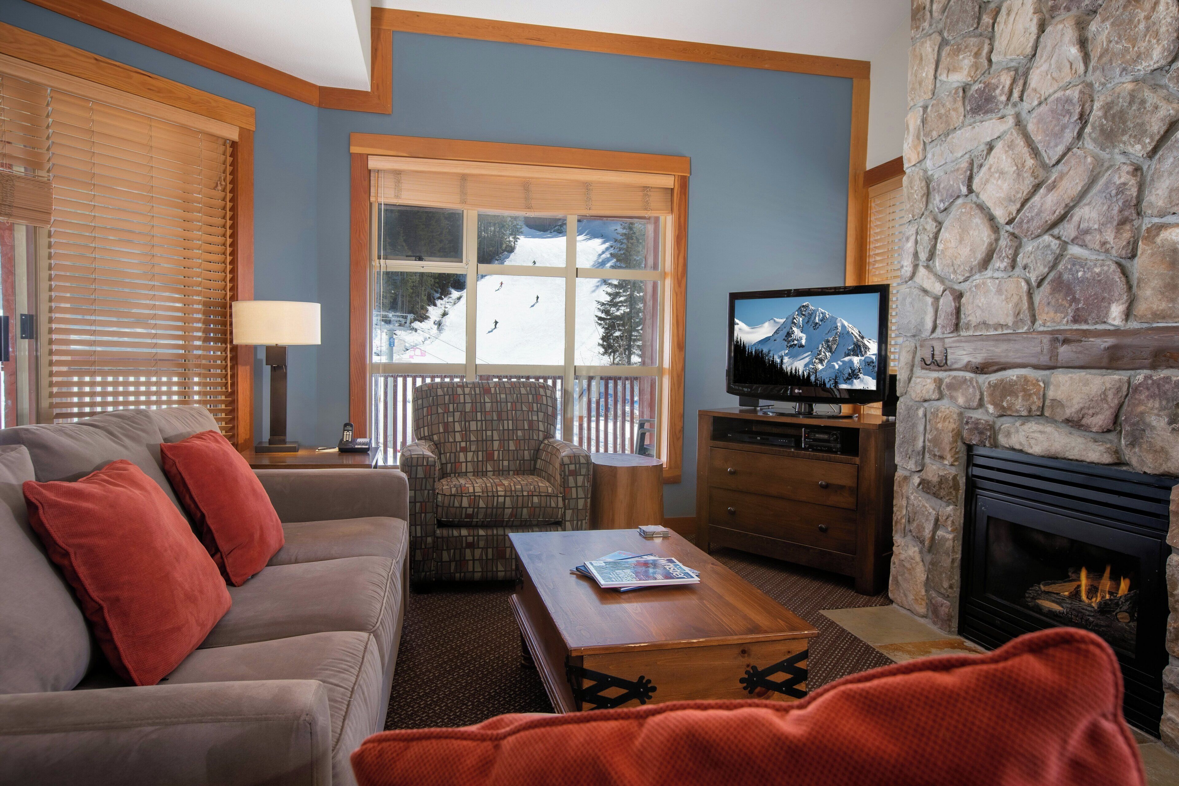 Una sala de estar con sofá, sillón, mesa de centro, chimenea, televisión y ventana con vistas a un paisaje nevado.
