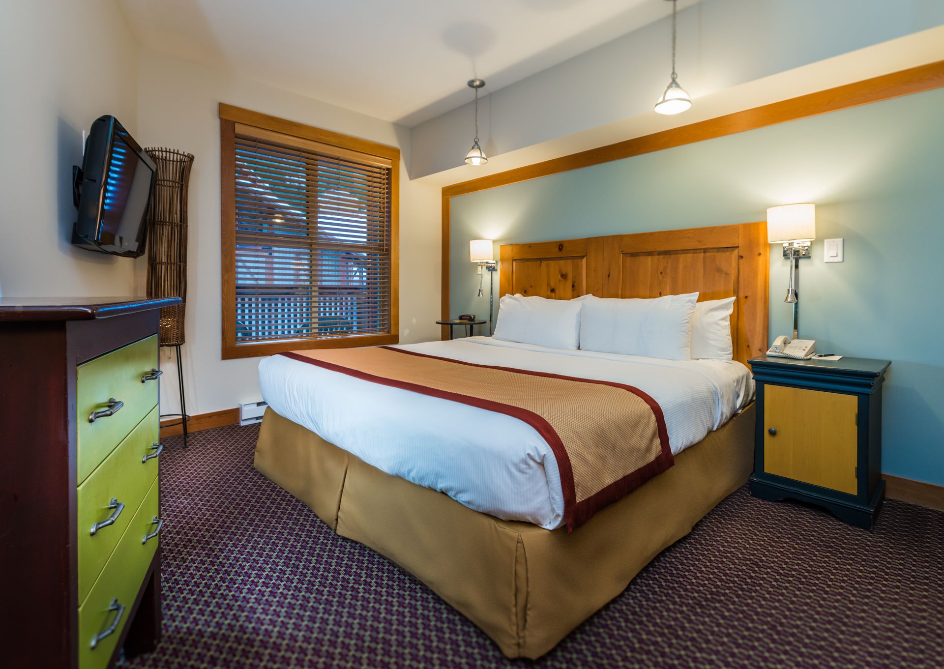 Ein Hotelzimmer mit einem großen Bett, einem Holzkopfende, zwei Nachttischlampen, einem Schreibtisch mit Telefon und einem an der Wand befestigten Fernseher.