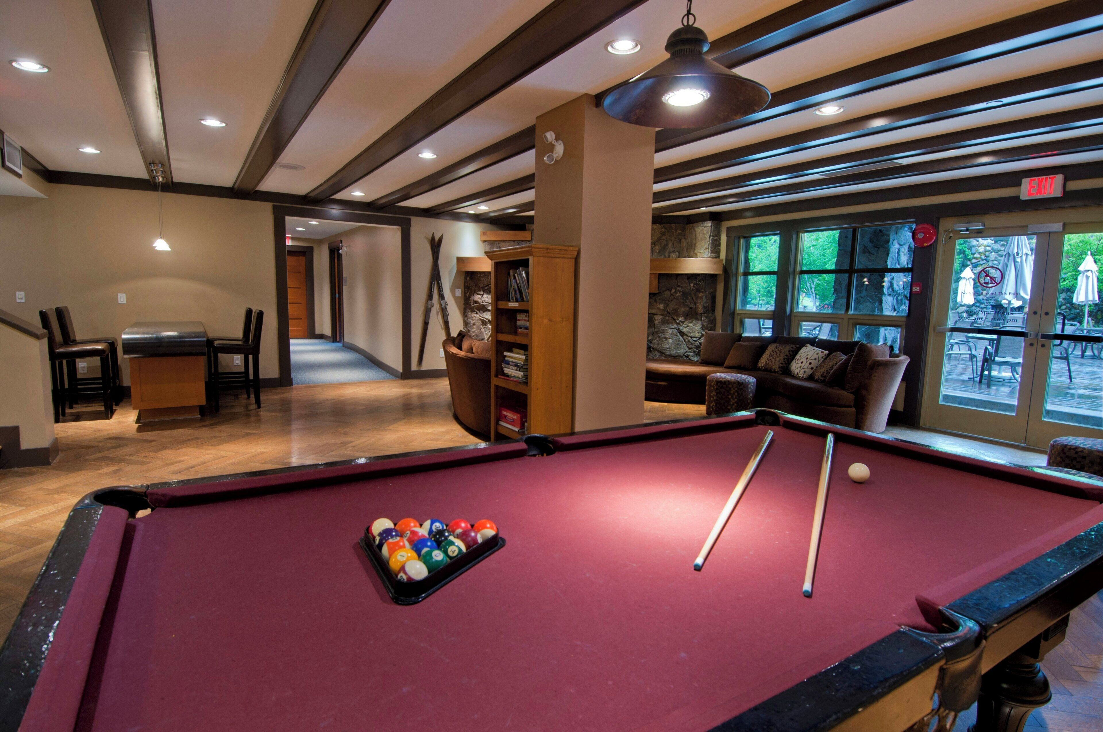 A pool table with balls, a sofa, and a bookshelf in a room with wooden beams and large windows.