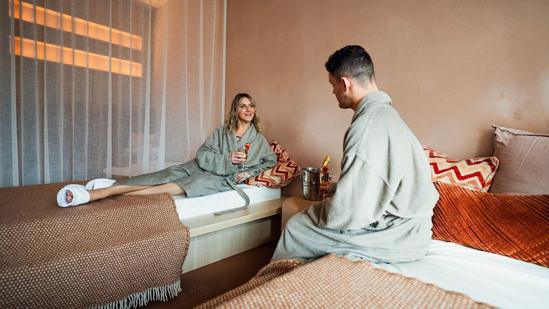 A hotel room with a bed, a bedside table with a bottle and glasses, and a window with sheer curtains.
