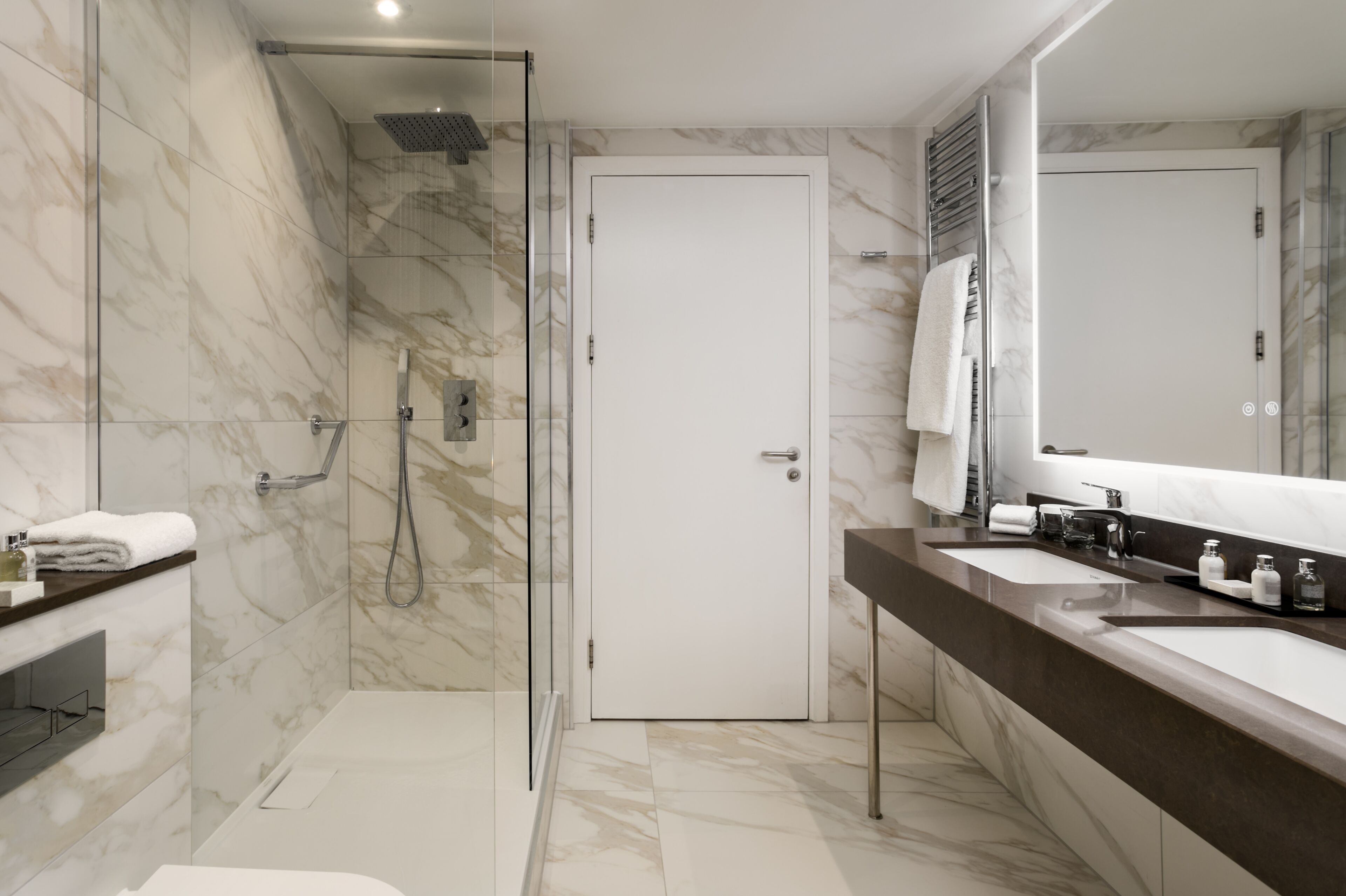 Een moderne badkamer met een douche met glazen wand, marmeren wanden en een grote spiegel boven de wastafel.