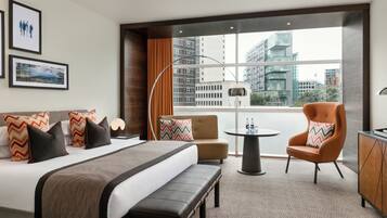 Habitación de hotel moderna con una cama grande, un sofá, una silla, una mesita y vistas a la ciudad a través de amplios ventanales.