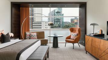 A modern hotel room with a large bed, a sofa, a chair, a small table, and a desk. The room has a large window with a city view.