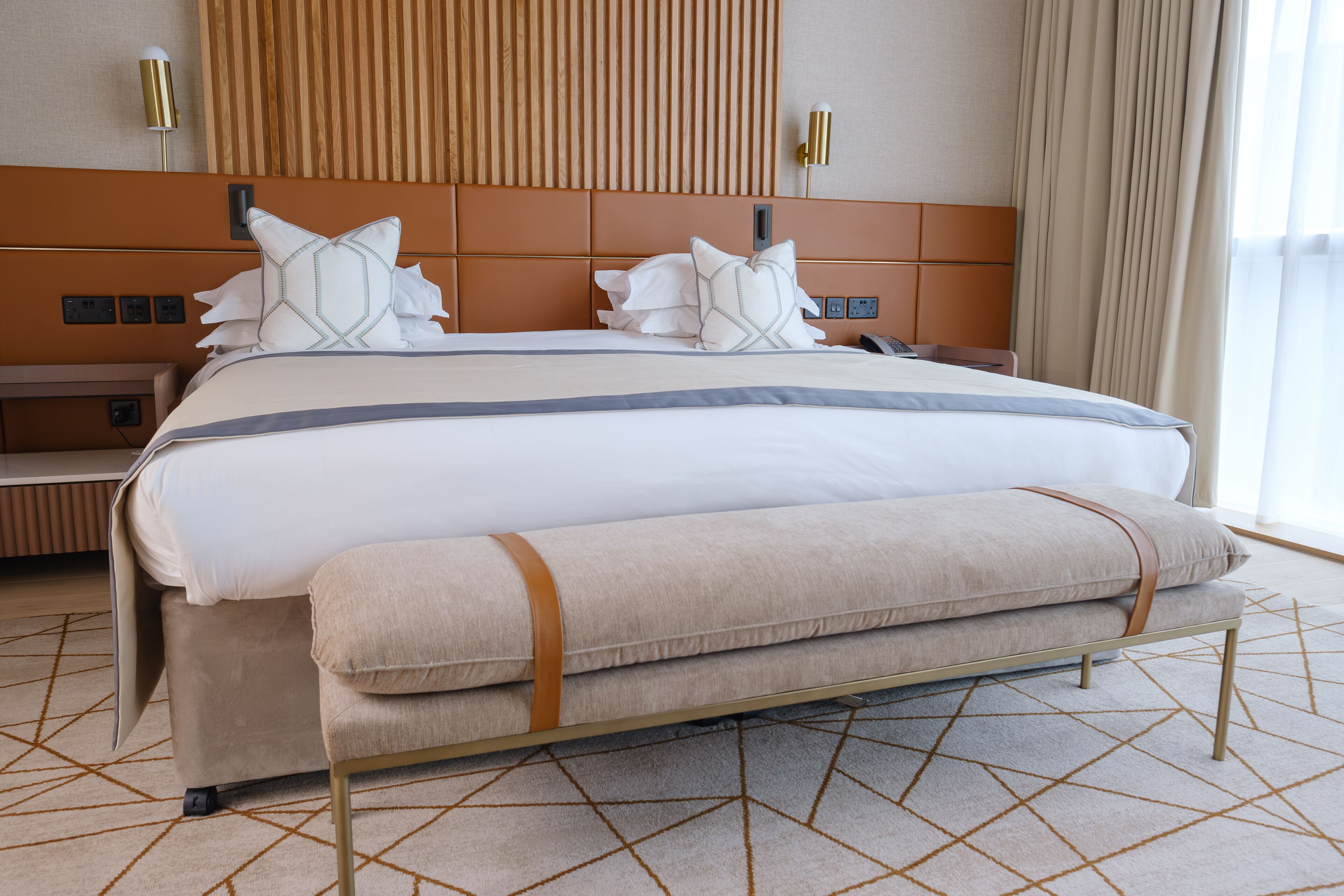 A modern hotel room with a large bed, a headboard with vertical slats, a bedside table, and a lamp.