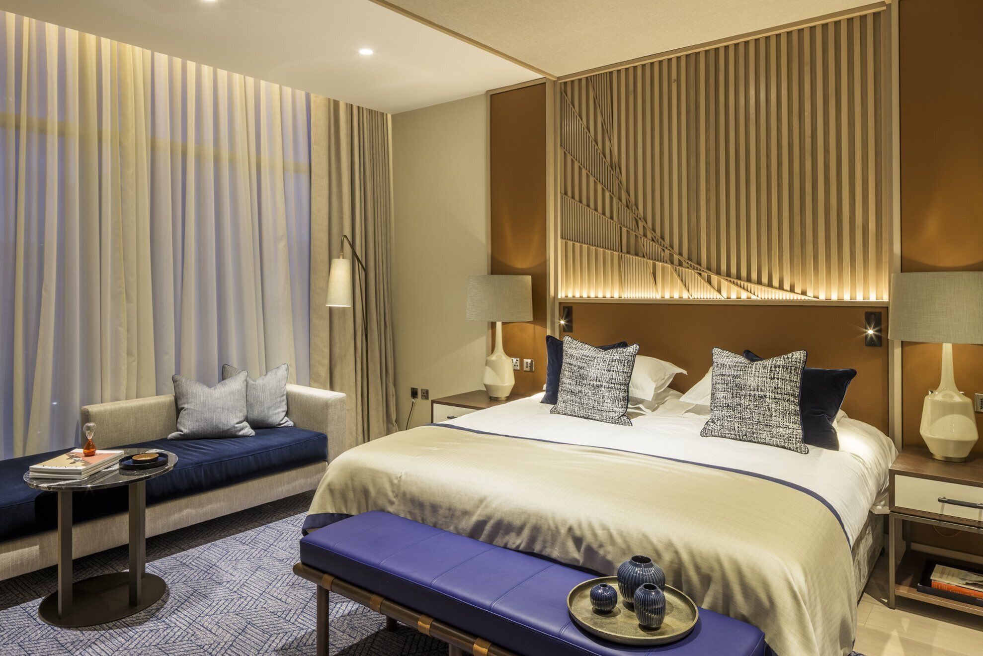 A modern hotel room with a large bed, a sofa, a small table, and a bench.