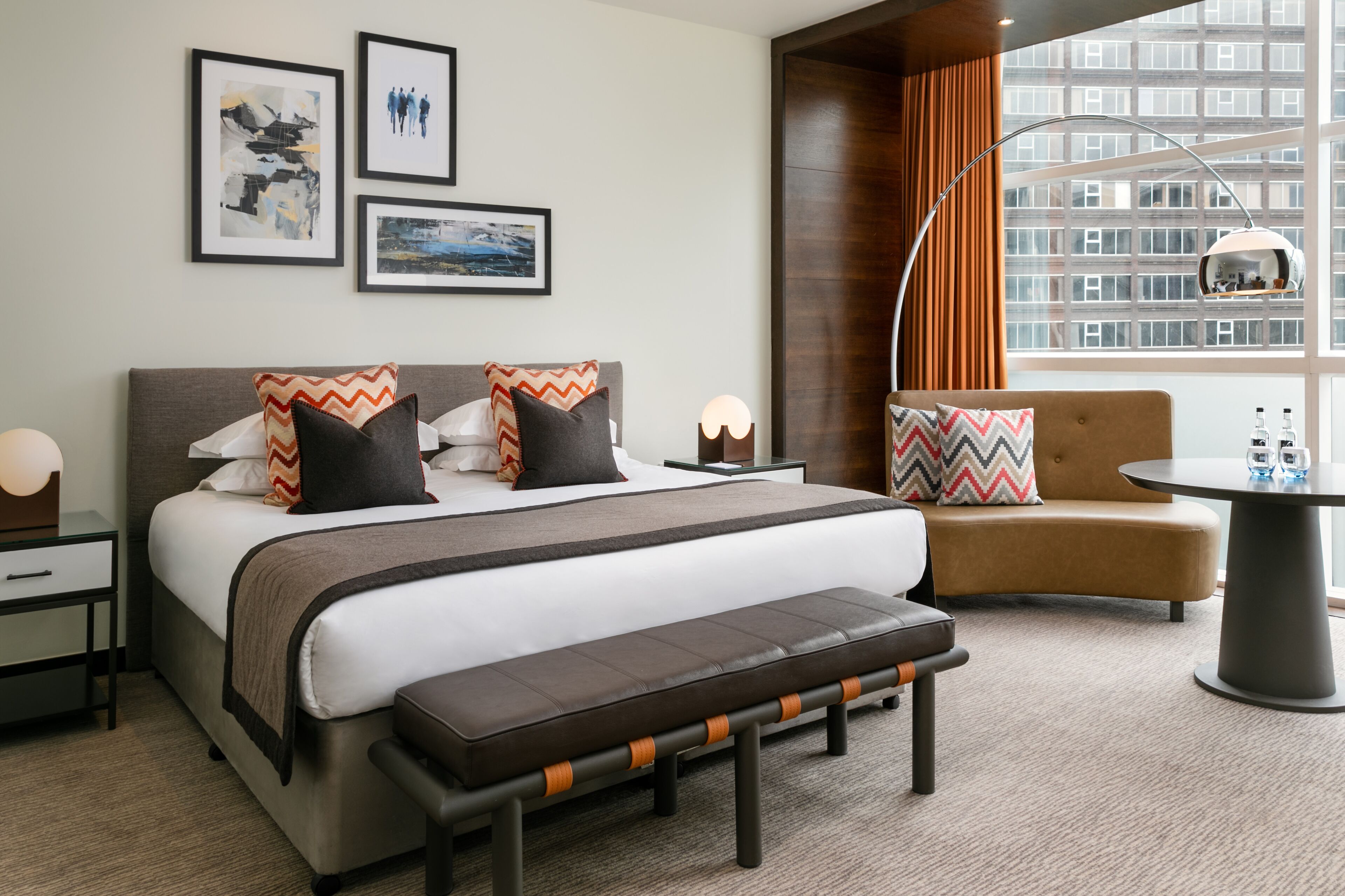 A modern hotel room with a large bed, a bench, a sofa, and a dining table. There are framed pictures on the wall and a view of buildings through the window.