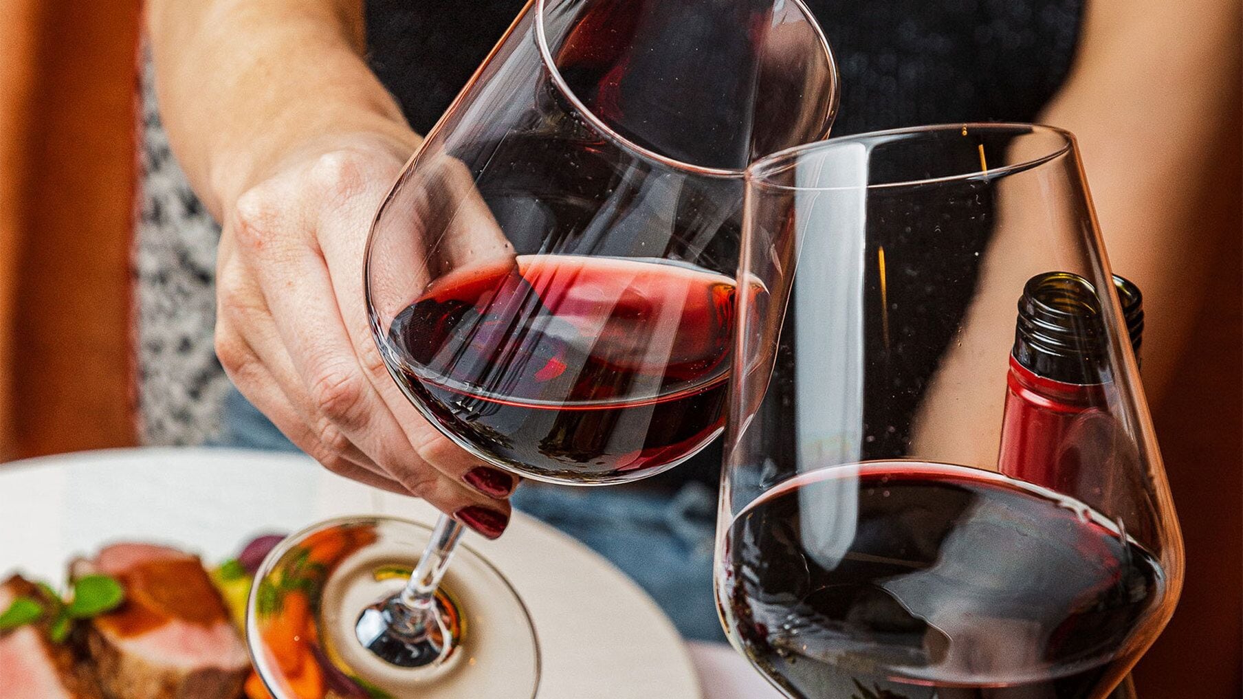 A person holding two wine glasses with a bottle of wine on the table.