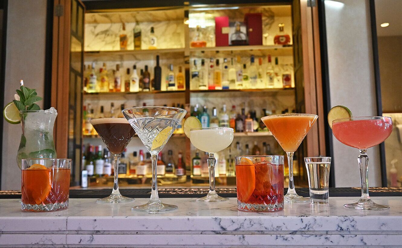Eine Bar mit verschiedenen Cocktails in unterschiedlichen Gläsern, einer Limettengarnitur und einer Auswahl an Spirituosenflaschen im Hintergrund.
