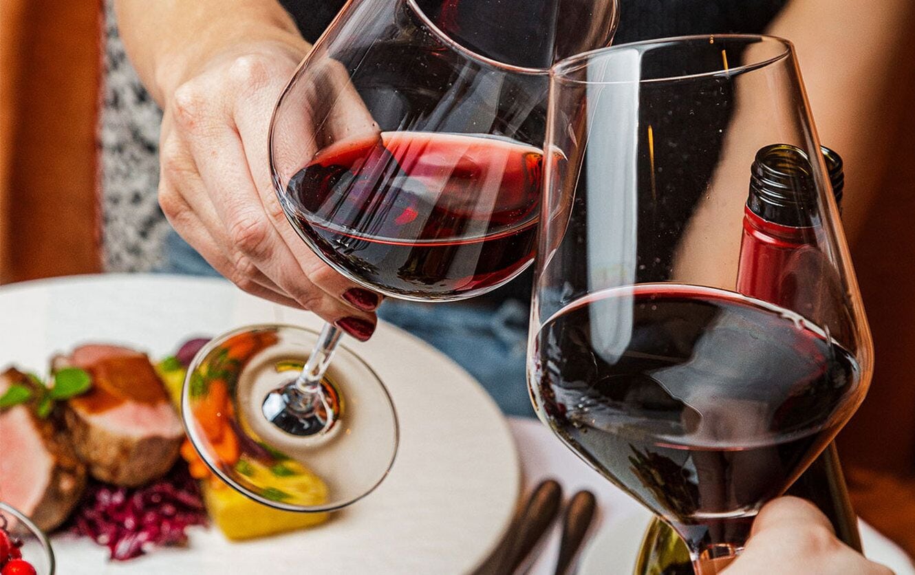 Una persona sosteniendo una copa de vino con líquido rojo, una botella de vino y un plato con comida.