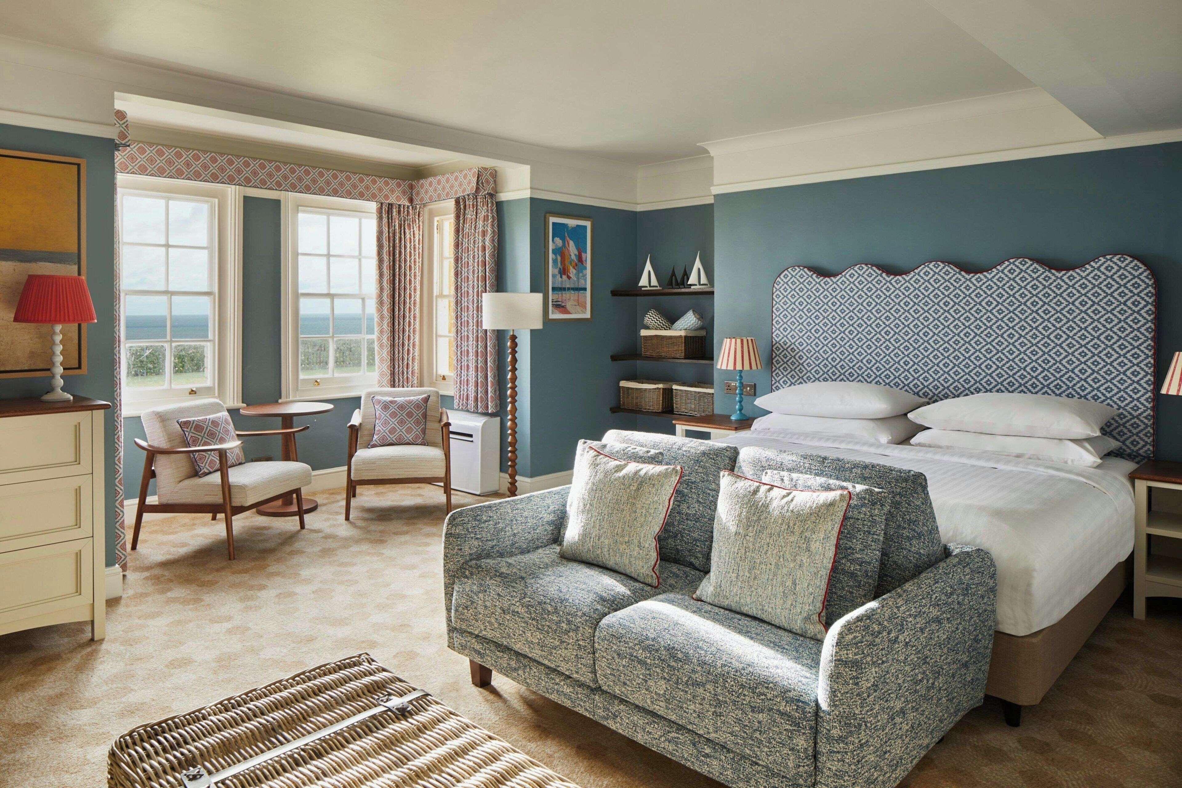 A hotel room with a bed, a sofa, a desk, and a chair. There is a window with curtains, a lamp, and a painting on the wall.