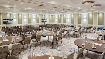 A large conference room with multiple tables and chairs, equipped with water bottles and glasses, and a television screen displaying a welcome message.
