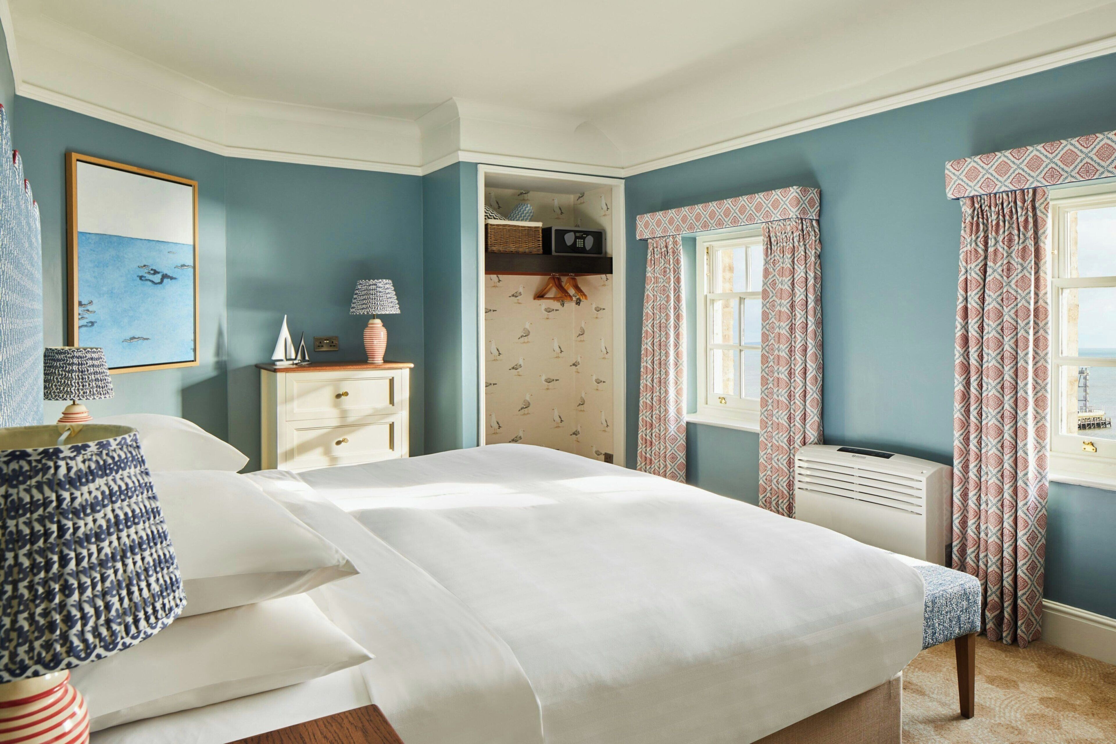 Un dormitorio con cama, mesitas de noche, lámpara, cómoda, ventana con cortinas y vista al mar.