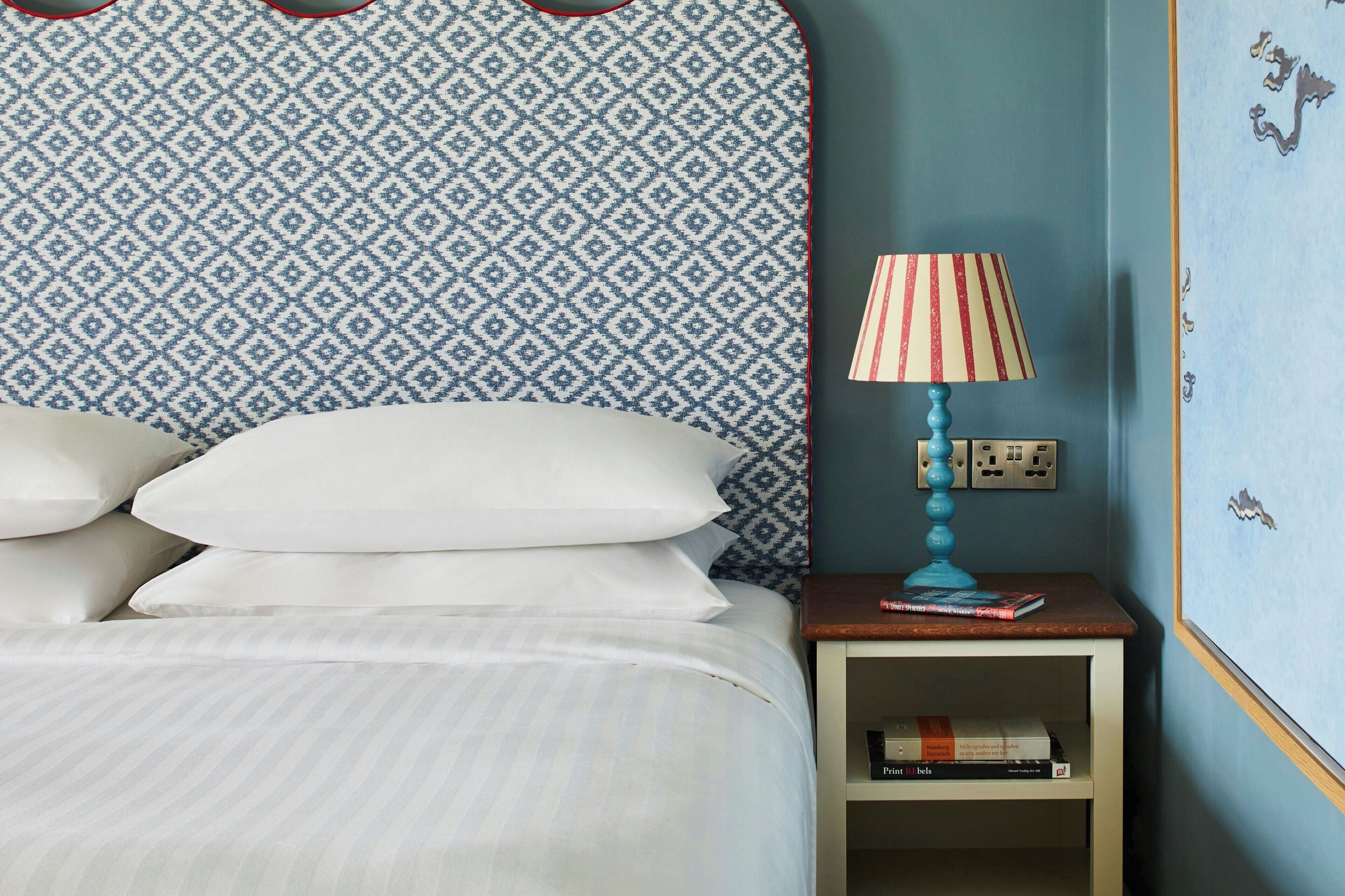 A neatly made bed with white pillows and a striped bedspread, a blue and white patterned headboard, a small bedside table with a lamp, and a wall with a blue paint job and decorative elements.