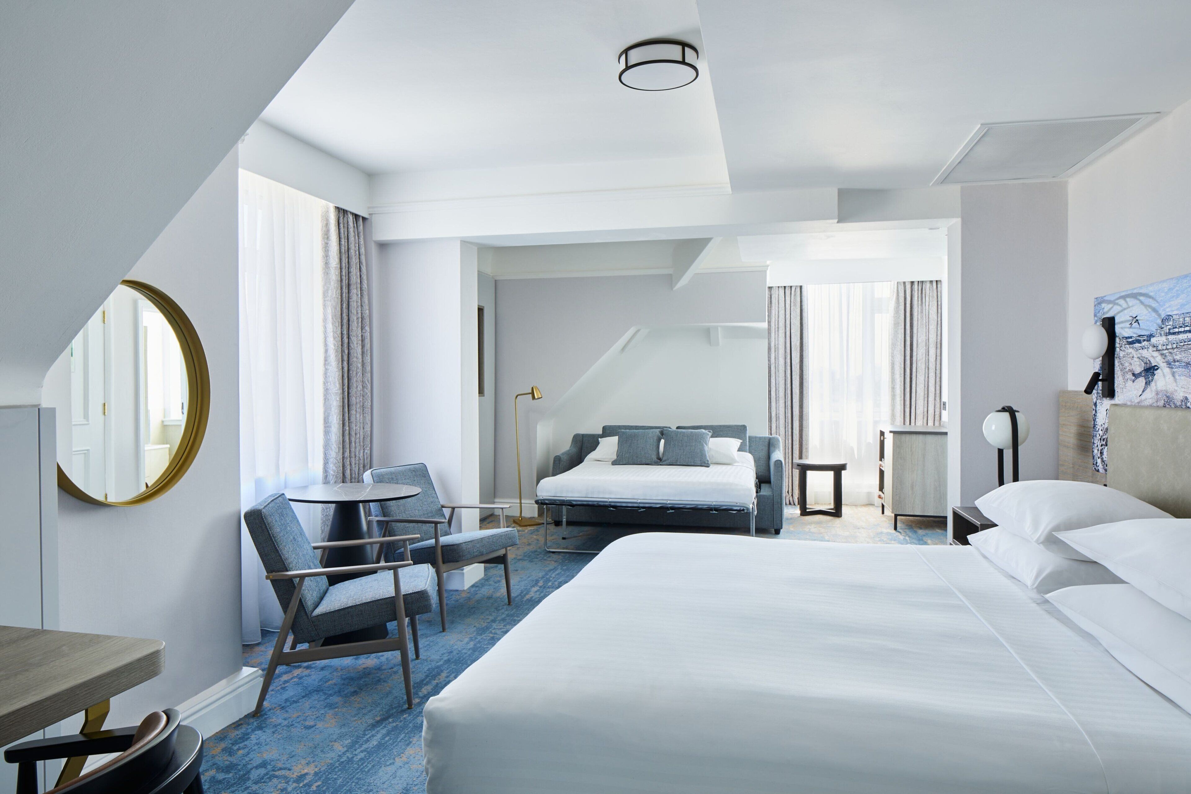 Une chambre d’hôtel avec un grand lit, un bureau, une chaise et un miroir rond.