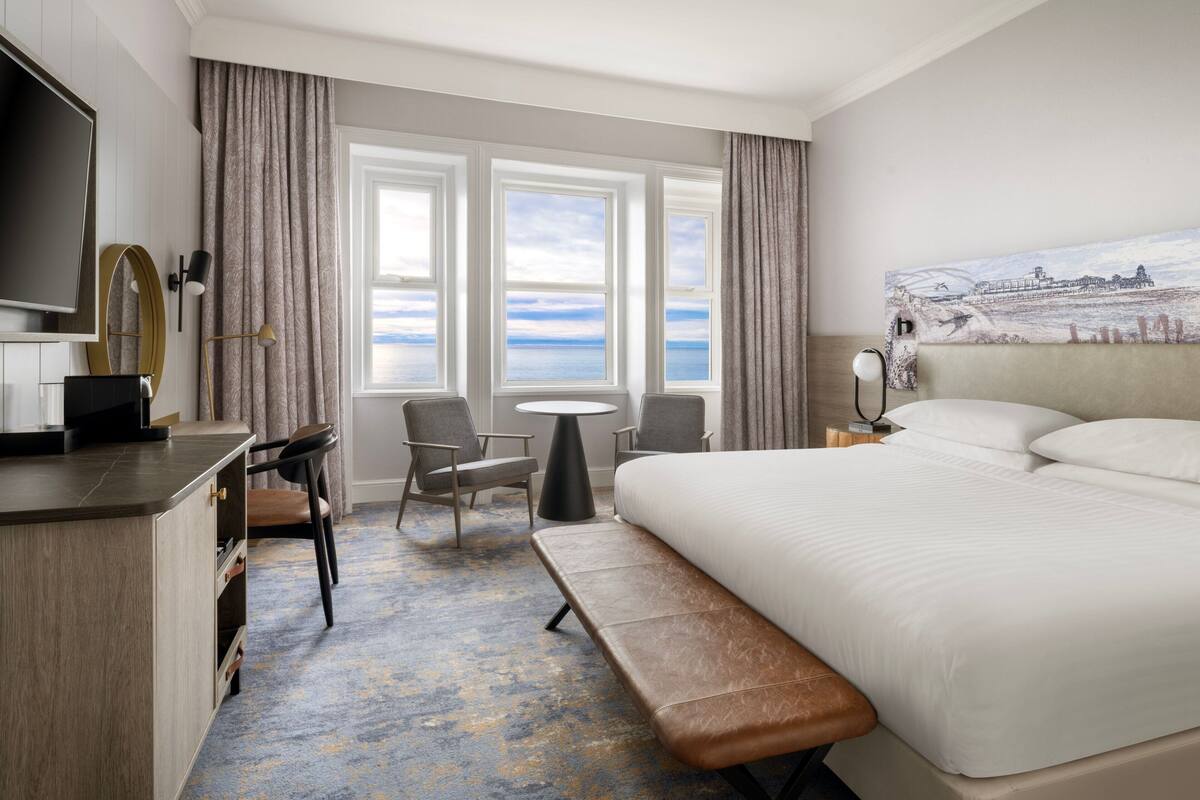 A hotel room with a large bed, a desk with a TV, a bench, two chairs, a small table, and a painting of a coastal scene.
