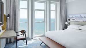 A hotel room with a large bed, a desk, and a chair. There are two windows with a view of the sea.