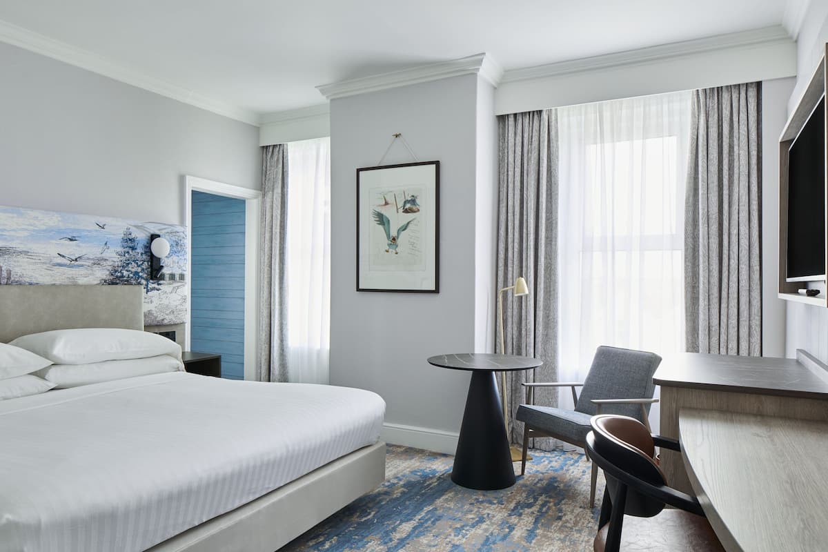 A hotel room with a large bed, a small round table, a chair, a desk, and a television.