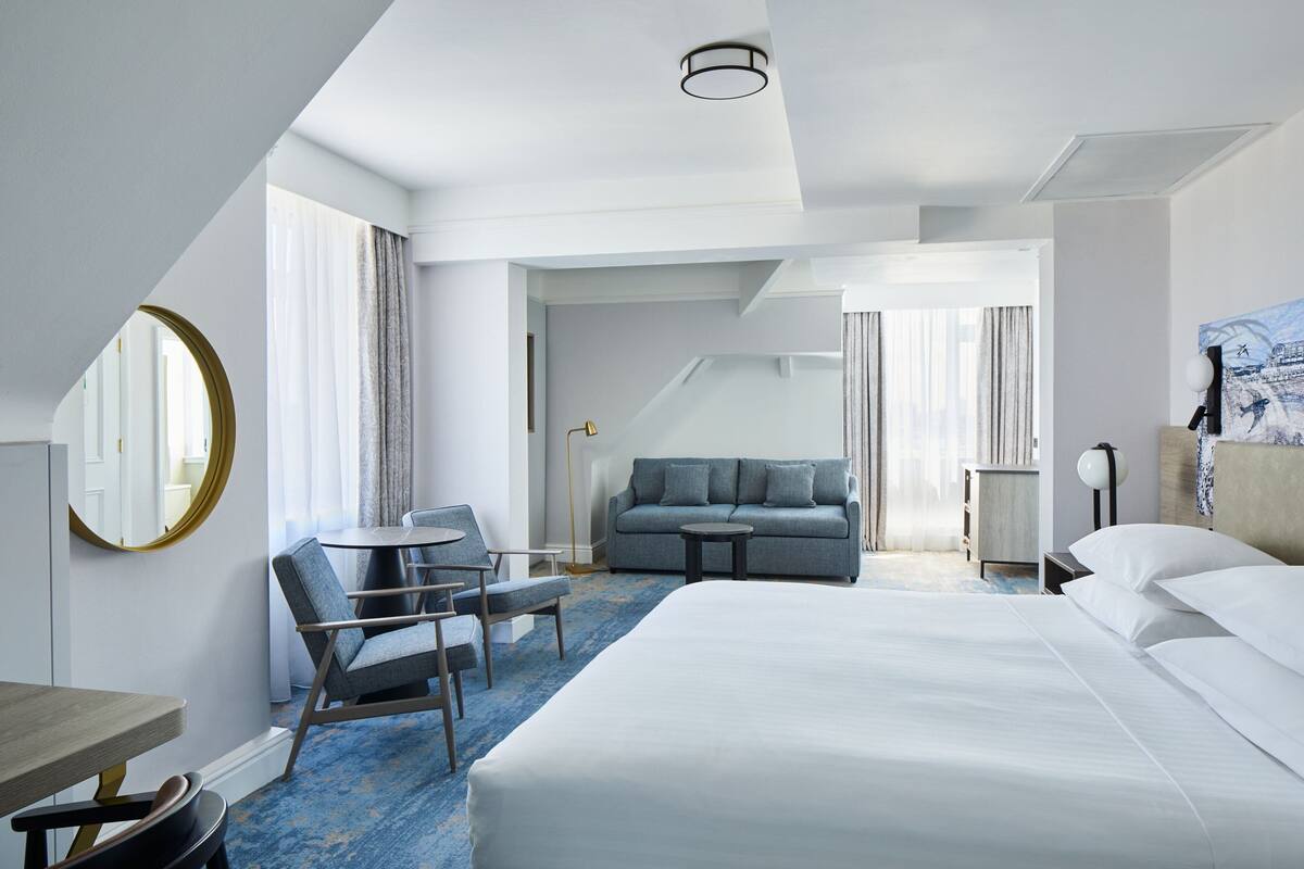 A hotel room with a large bed, a sofa, a dining table, and a chair. There is a round mirror on the wall and a painting on the side.