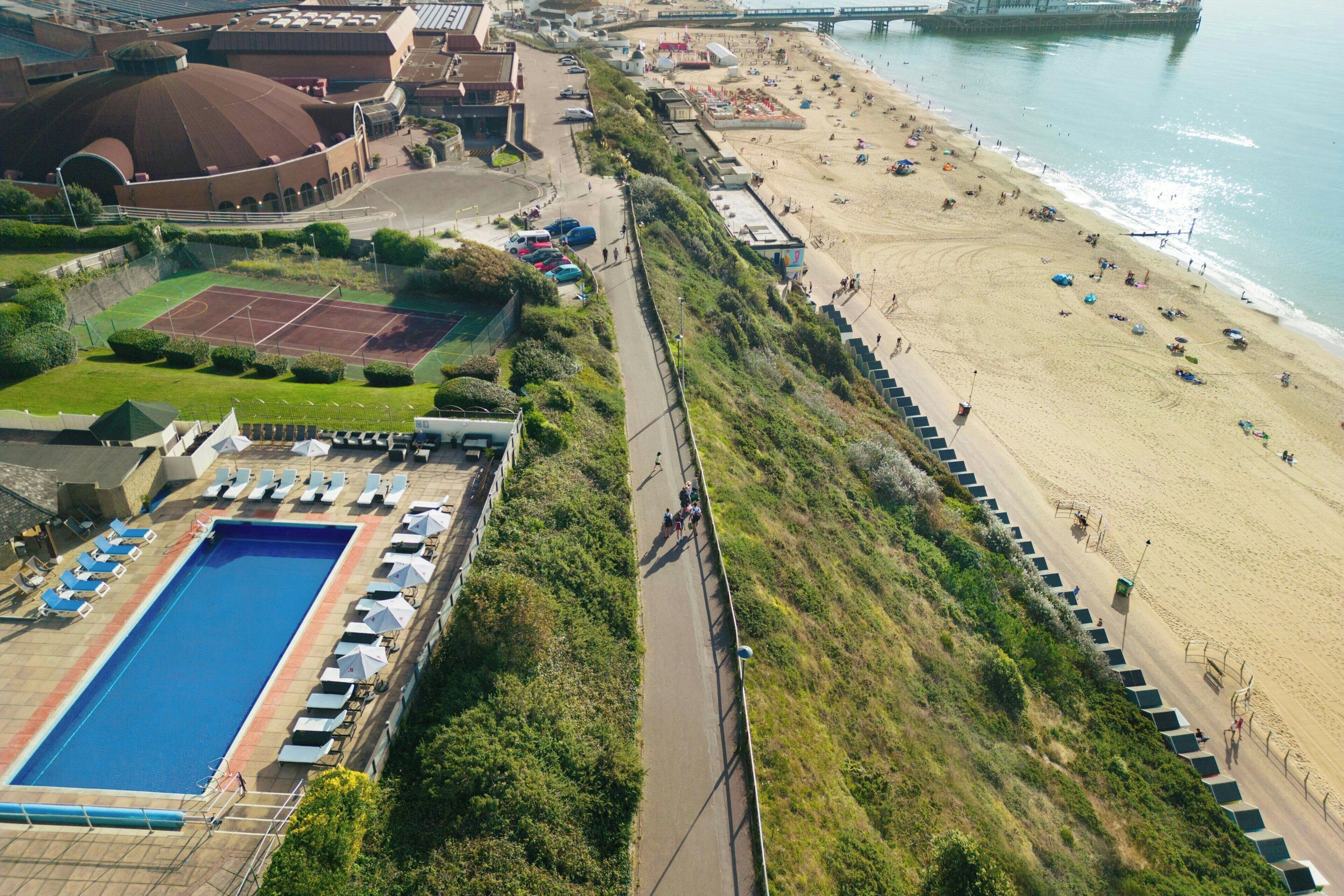 An aerial view of a coastal area with a swimming pool, beach, and a bridge in the background.